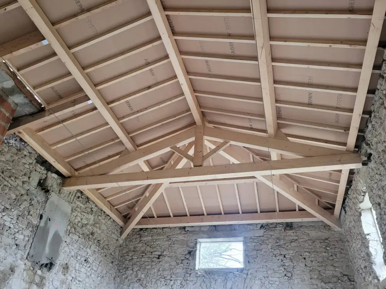 Vue en contre-plongée d'une charpente de toiture en bois, en cours d'installation, avec des panneaux de contreplaqué fixés.
