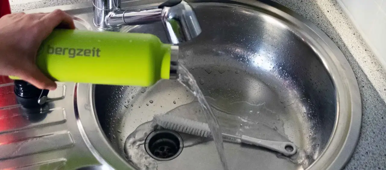 Eine Hand hält eine grüne Thermoskanne über ein Spülbecken, während Wasser hineinfließt. Eine Bürste liegt im Becken, bereit zur Thermoskanne reinigen.
