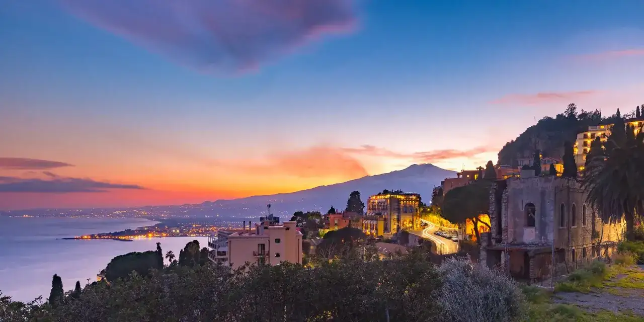 Tramonto su Taormina con l'Etna sullo sfondo. La costa illuminata si estende verso il mare calmo.