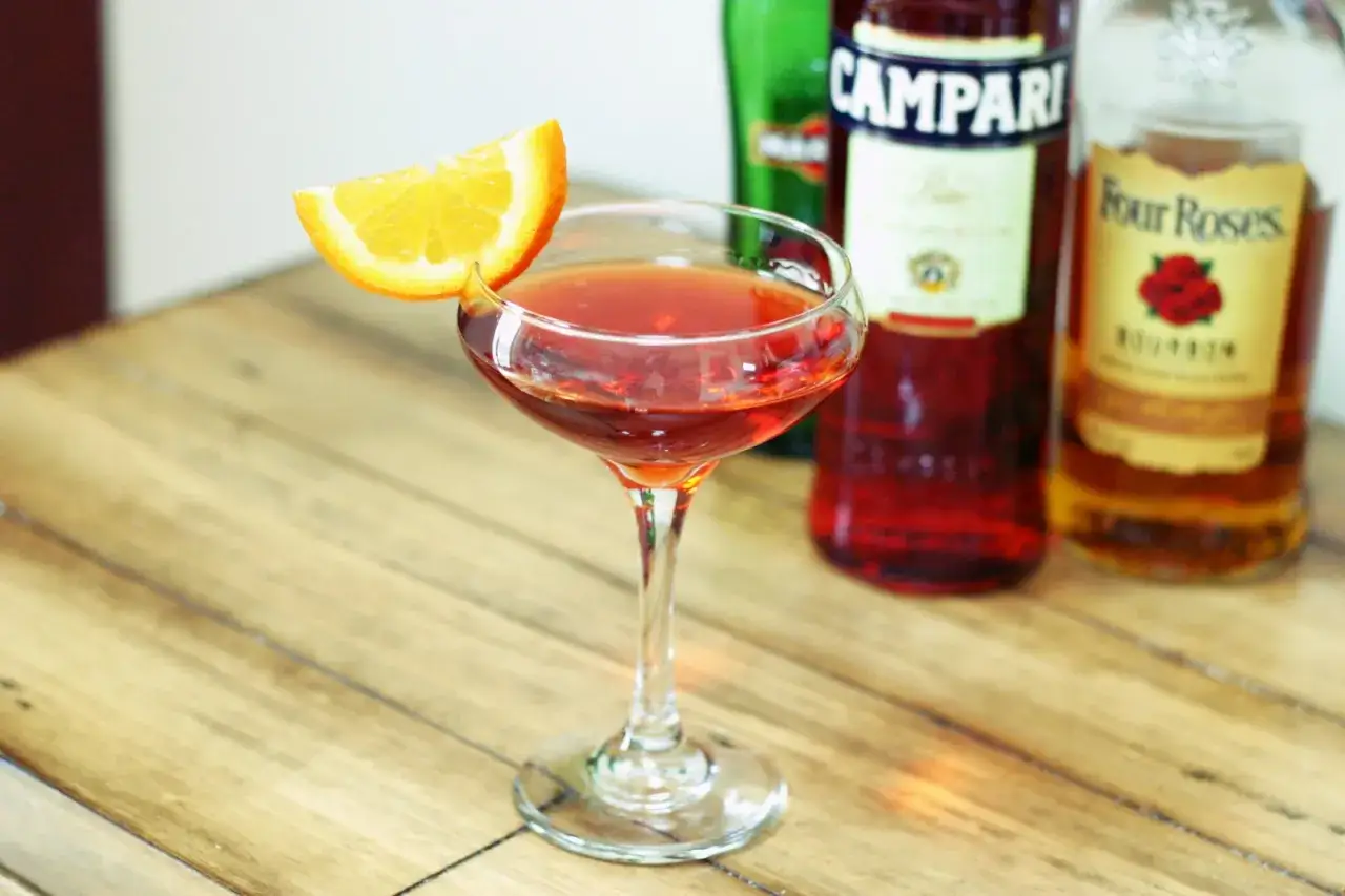 Elegancki boulevardier w kieliszku, ozdobiony plasterkiem pomarańczy. Obok butelki Campari i bourbonu Four Roses.