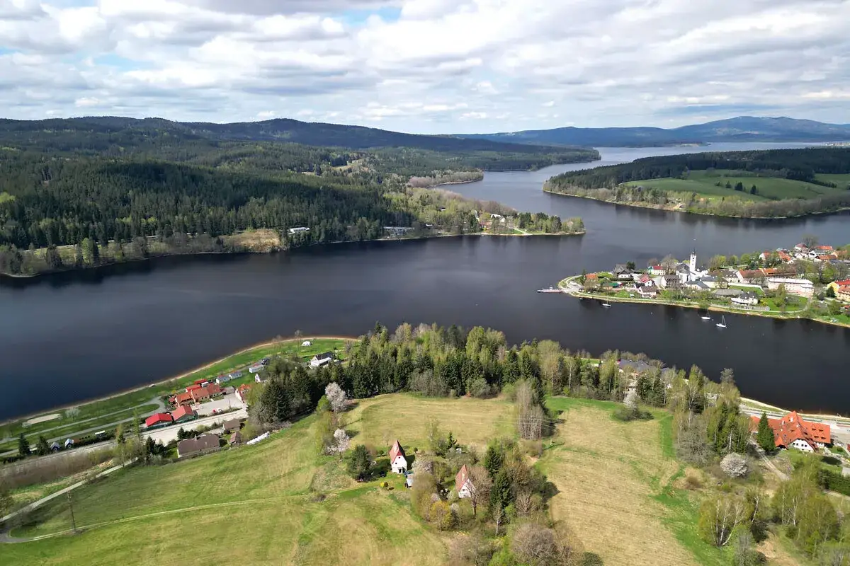 Blick &uuml;ber den Lipno Stausee mit bewaldeten H&uuml;geln, einer kleinen Stadt und gr&uuml;nen Wiesen.