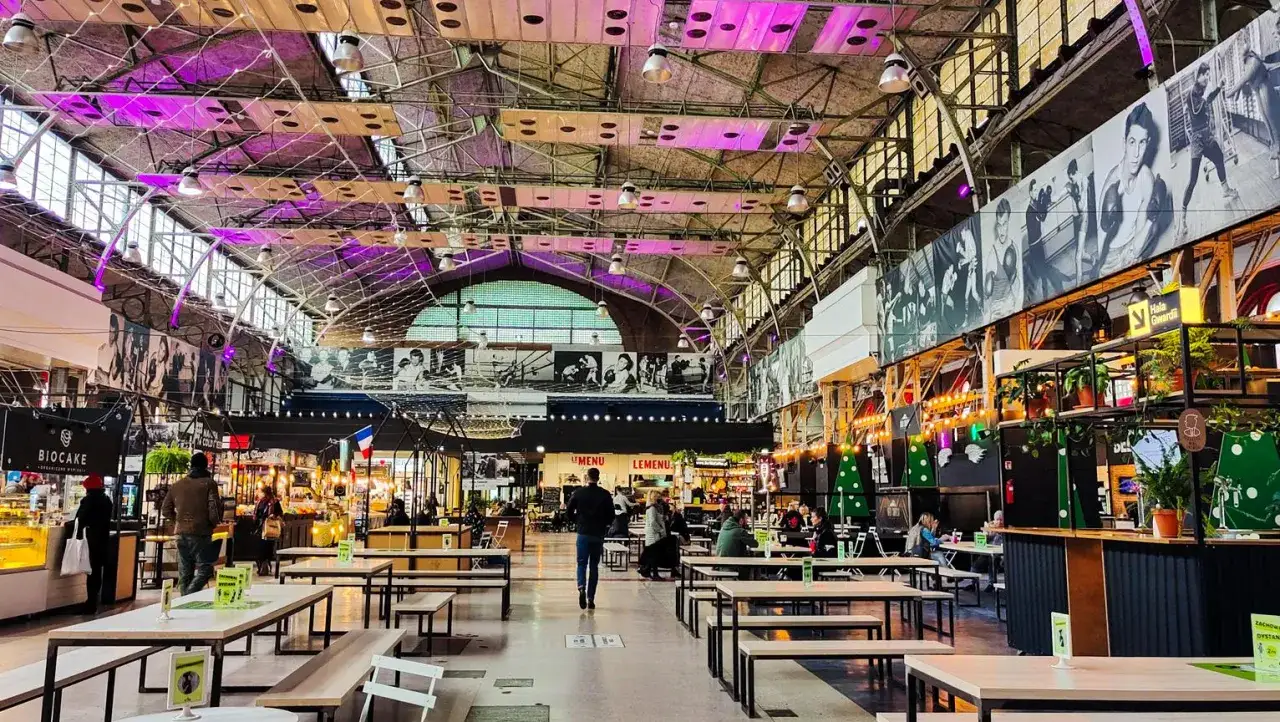 Wnętrze hali z licznymi stolikami i ławkami, gdzie znajduje się vege restauracja w Warszawie. W tle widać stoiska z jedzeniem i ludzi.