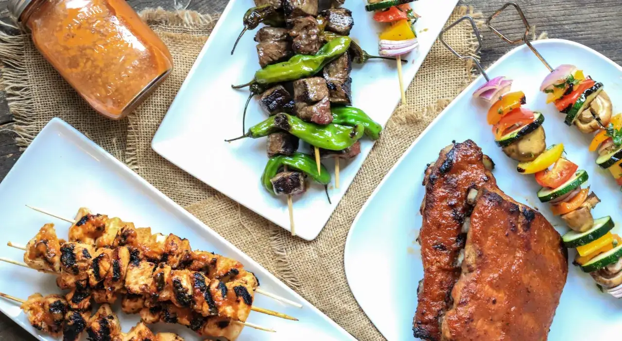 En burk med sås till grillat bredvid grillspett med kött, kyckling och grönsaker.