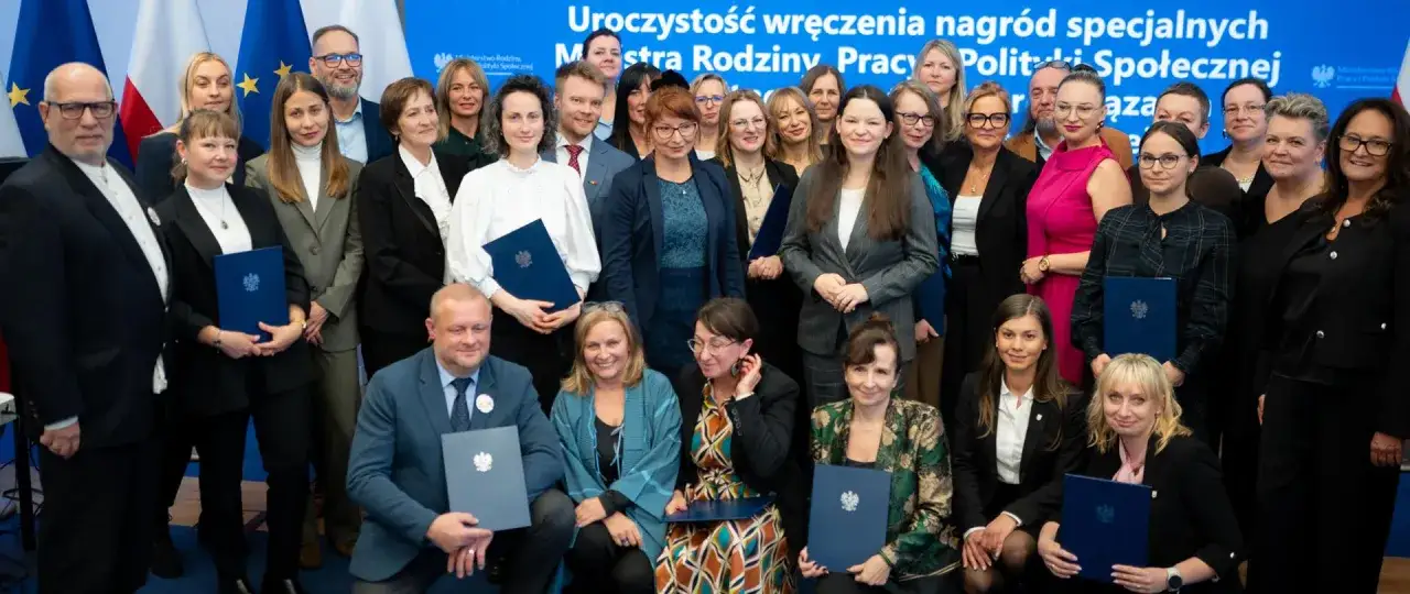 Grupa osób, w tym pracownik socjalny, odbiera nagrody. Wszyscy pozują do zdjęcia z dyplomami.