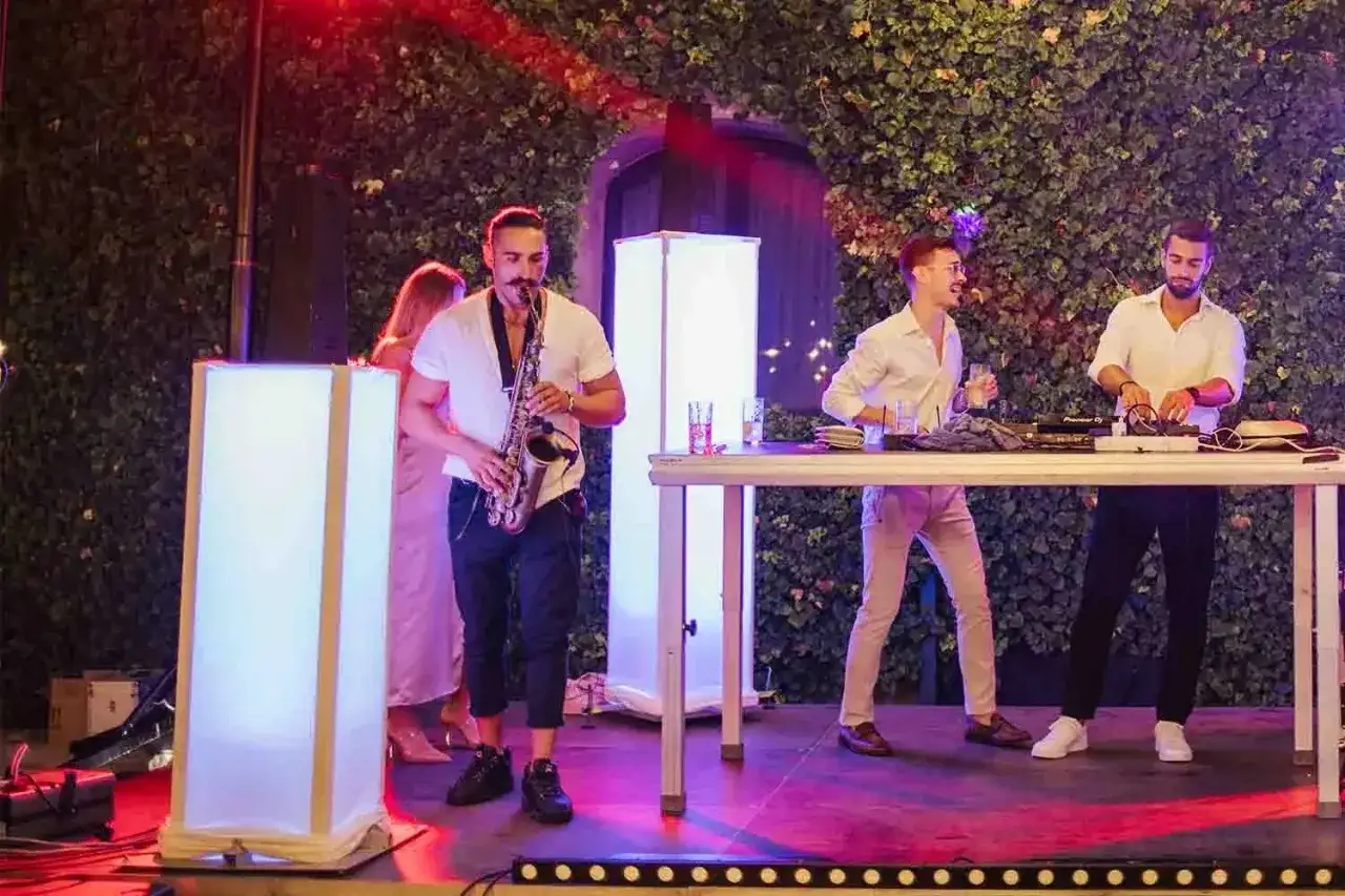 Un dj sax suona dal vivo mentre due dj mixano musica. Luci colorate illuminano la scena.