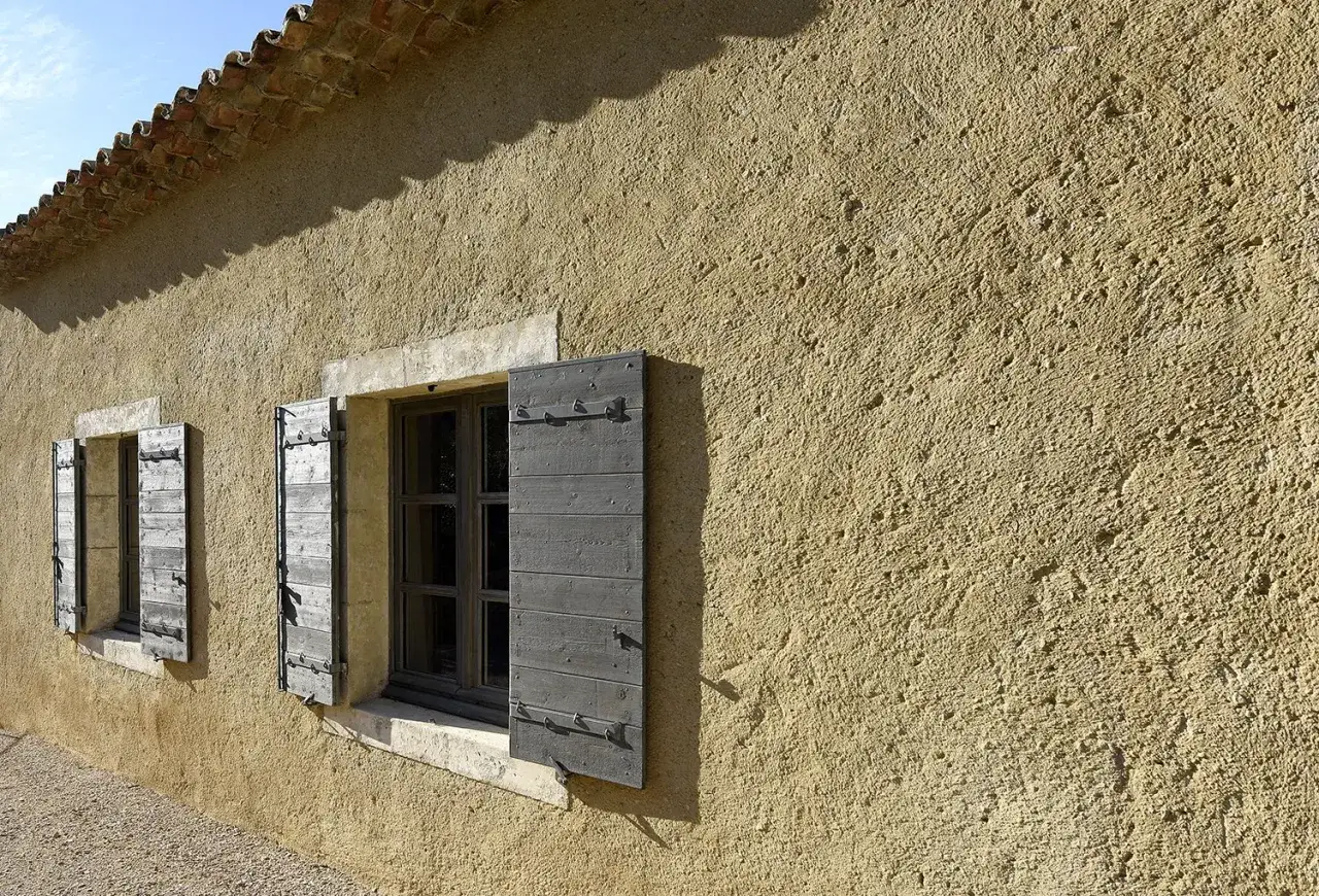 Fa&ccedil;ade d'une maison avec un enduit &agrave; la chaux ext&eacute;rieur textur&eacute;, deux fen&ecirc;tres aux volets en bois gris ouverts, et un toit de tuiles romanes.