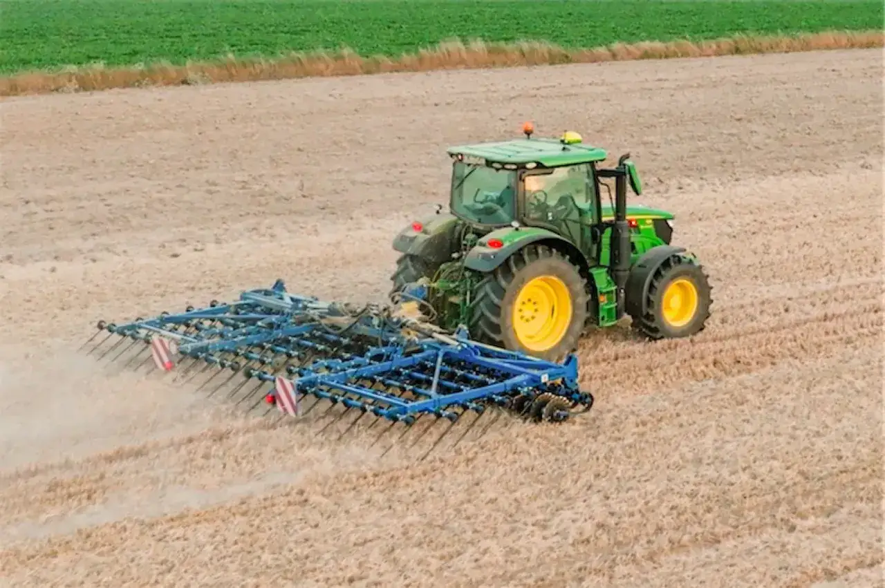 Zielony traktor z agregatem uprawowym, niczym buster, spulchnia ziemię na polu.