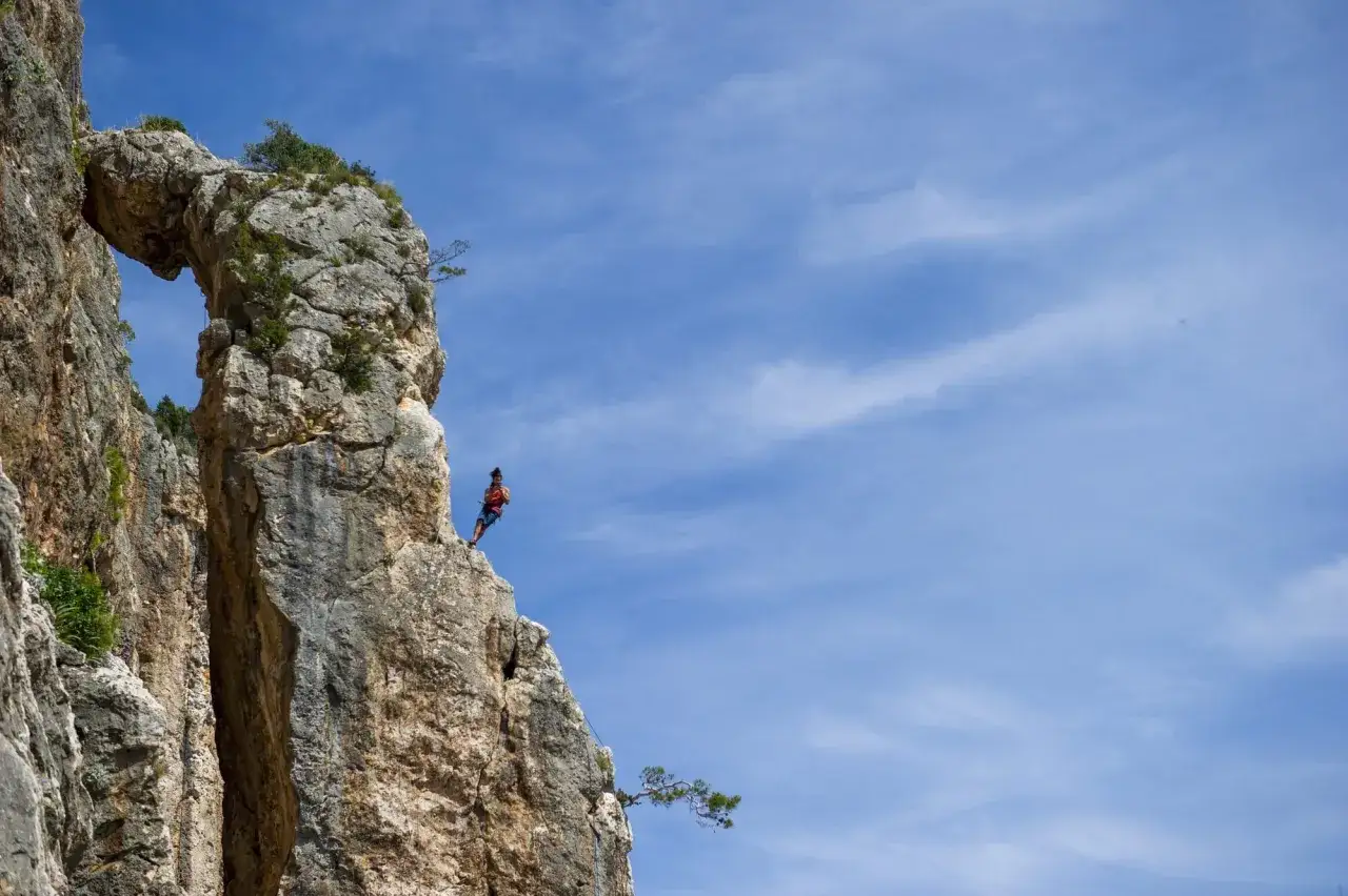Ein Kletterer genie&szlig;t die Aussicht beim Klettern in Kroatien, gesichert durch ein Seil an einer Felsformation mit einem nat&uuml;rlichen Bogen.