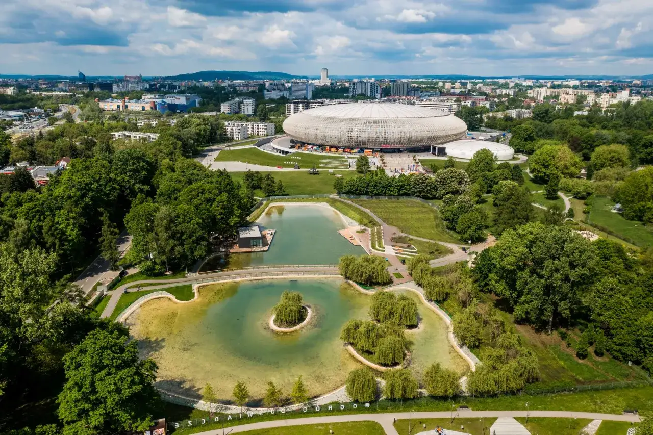 Malowniczy park lotnik&oacute;w polskich z jeziorem i wyspą, w tle hala widowiskowa i miasto.
