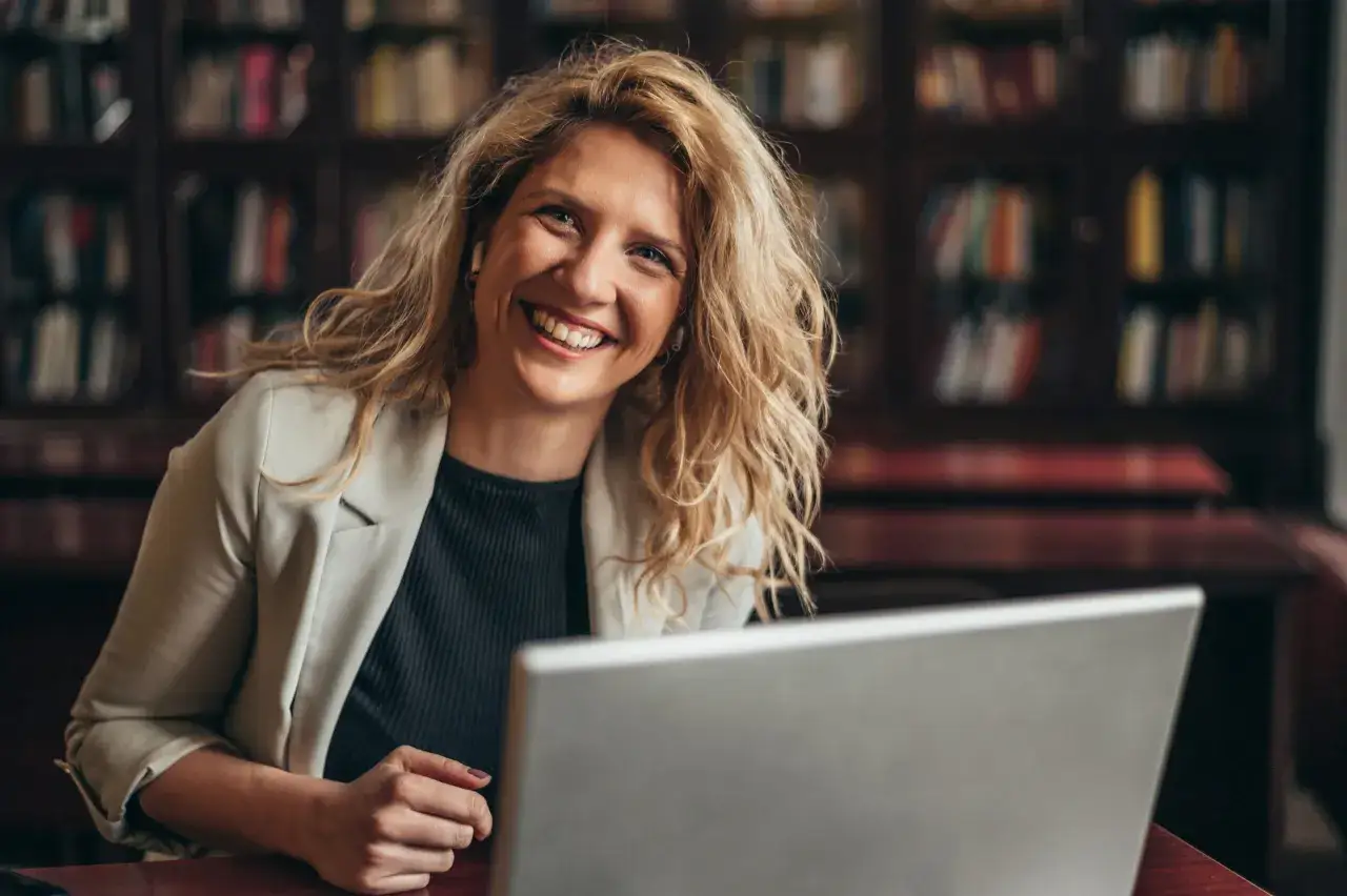 L&auml;chelnde Frau mit blonden Haaren und Kopfh&ouml;rern sitzt vor einem Laptop in einer Bibliothek. Sie vertieft sich in ihr gerontologie studium.