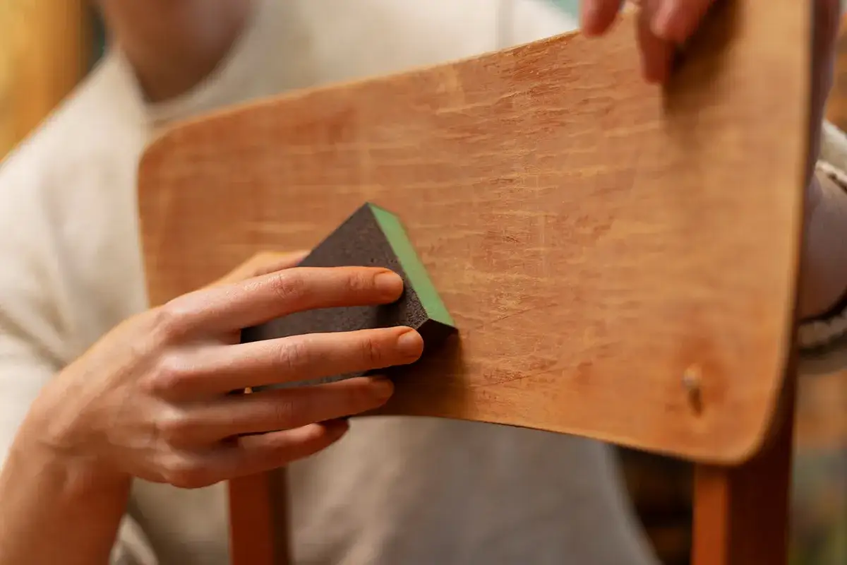 Mani che levigano un pezzo di legno con una spugna abrasiva, un passo fondamentale per imparare come lucidare il legno e ridargli vita.