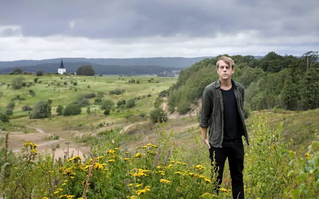 Christoffer Carlsson står i en blomsteräng med en kyrka i bakgrunden. Landskapet påminner om hans barndomshem, en plats som ofta inspirerar hans berättelser om familj.
