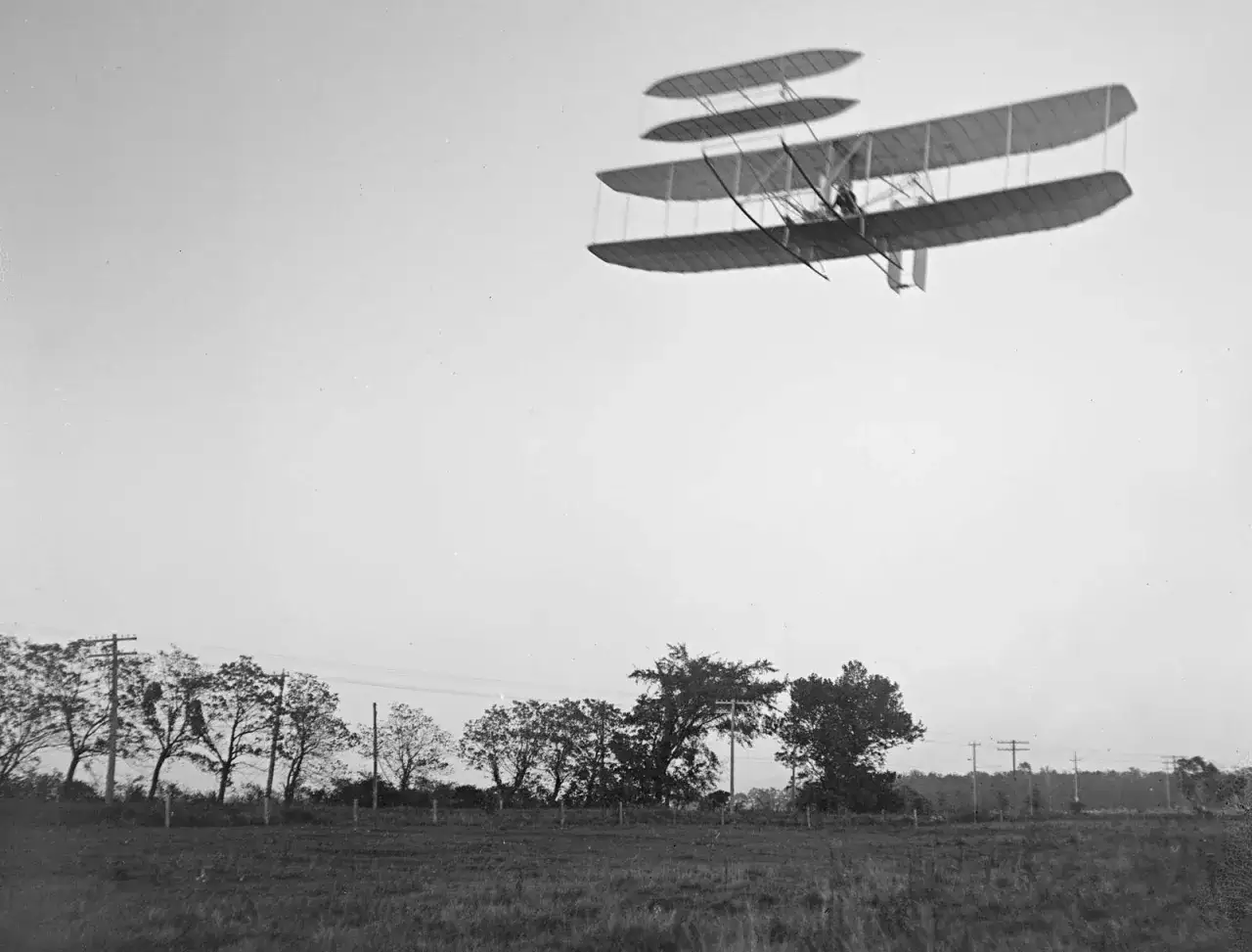 Der Wright Flyer fliegt über ein Feld. Bäume und Strommasten säumen den Horizont.