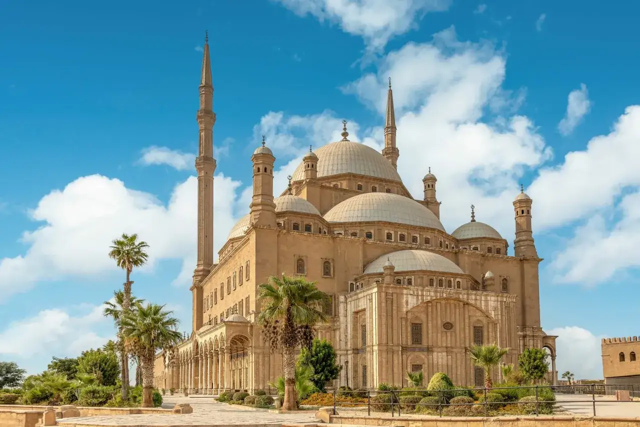 Die Moschee von Muhammad Ali Pascha, ein Wahrzeichen der cairo stadt, ragt majestätisch unter blauem Himmel mit Wolken auf. Palmen säumen den Weg.