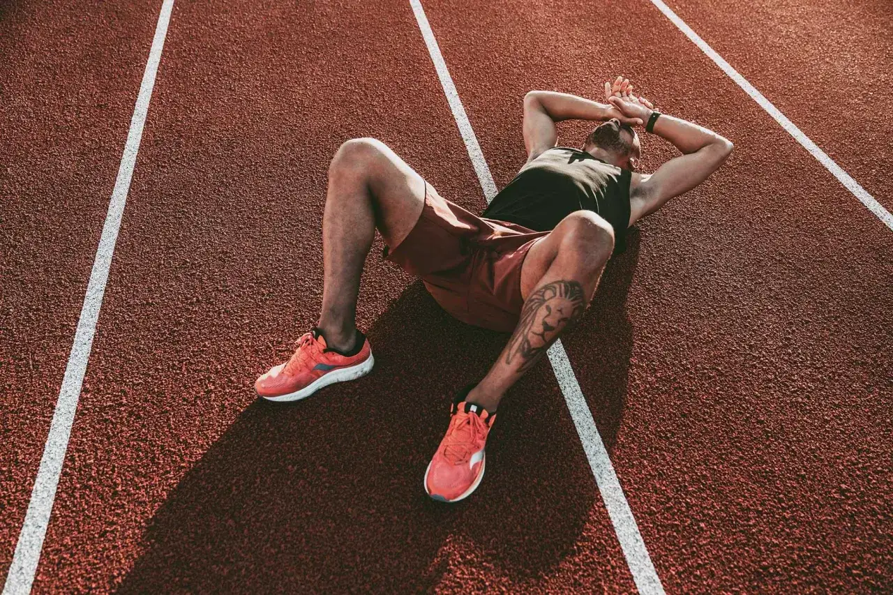 Atleta esausto a terra su una pista da corsa, mani sulla testa, possibili sintomi di overtraining.
