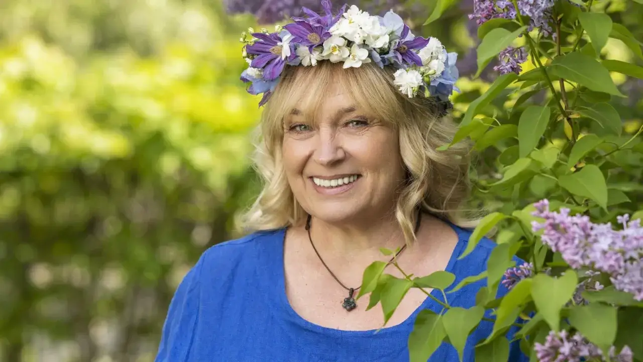 Sussie Eriksson med blomsterkrans i lila och vita blommor, klädd i blått, omgiven av syren. Känd från filmer och tv-program.