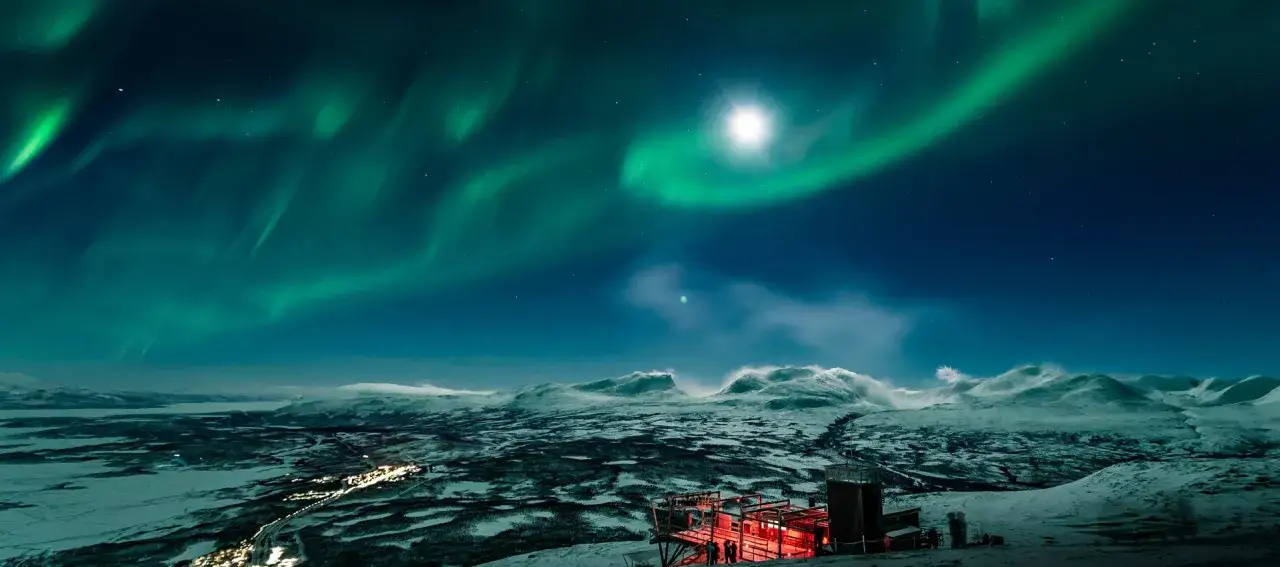 Norrsken dansar över ett snötäckt landskap. En upplyst byggnad med röda lampor står på en kulle, med en **arctic road** som slingrar sig genom dalen nedanför.