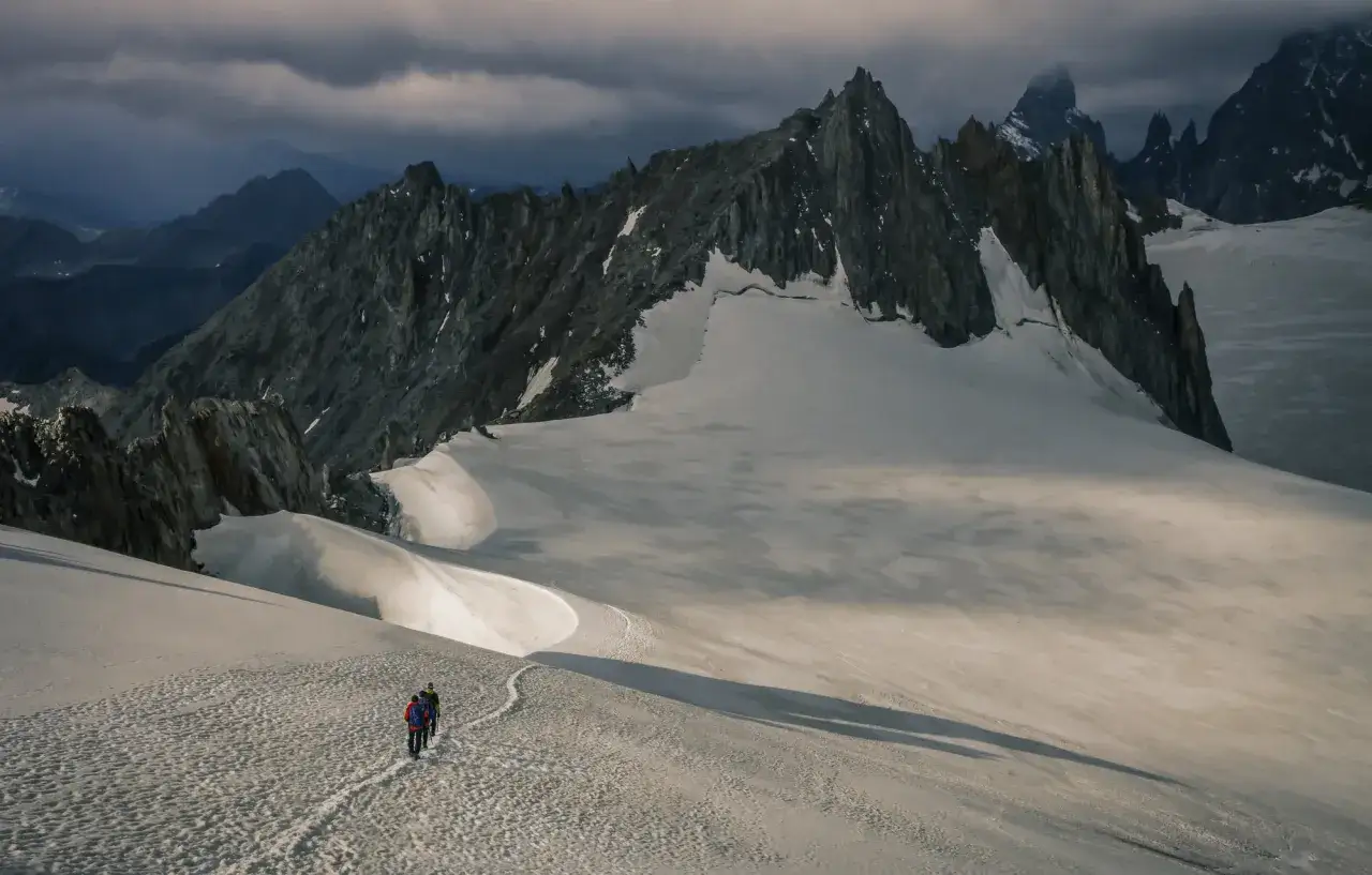 Deux randonneurs suivent un sentier sur un glacier, sous un ciel mena&ccedil;ant, dans un paysage de montagne spectaculaire. Un trail mythique en France.