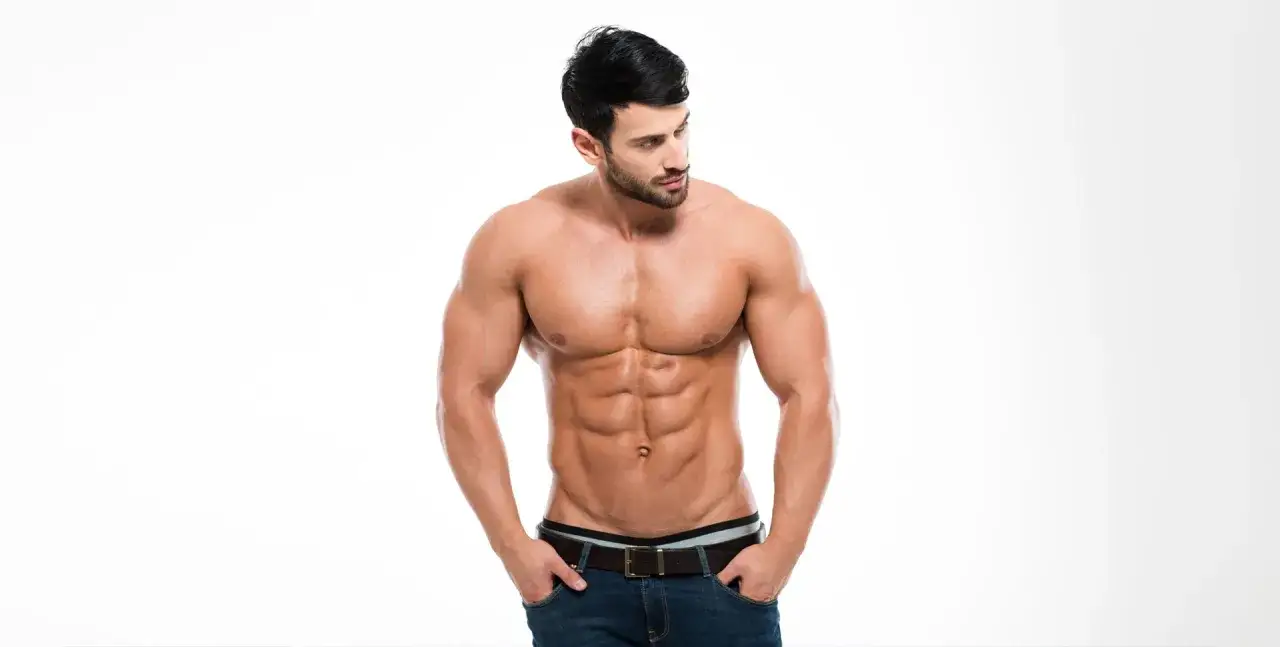 Uomo con addominali scolpiti e pettorali definiti, in jeans, guarda di lato.