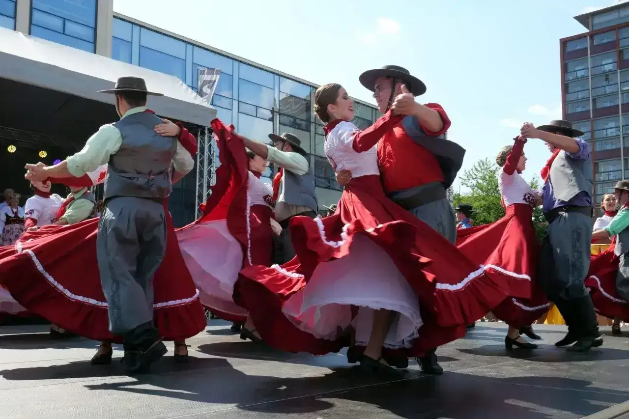 Tancerze w tradycyjnych strojach, z falującymi czerwonymi spódnicami, wykonują żywiołową polkę na scenie.