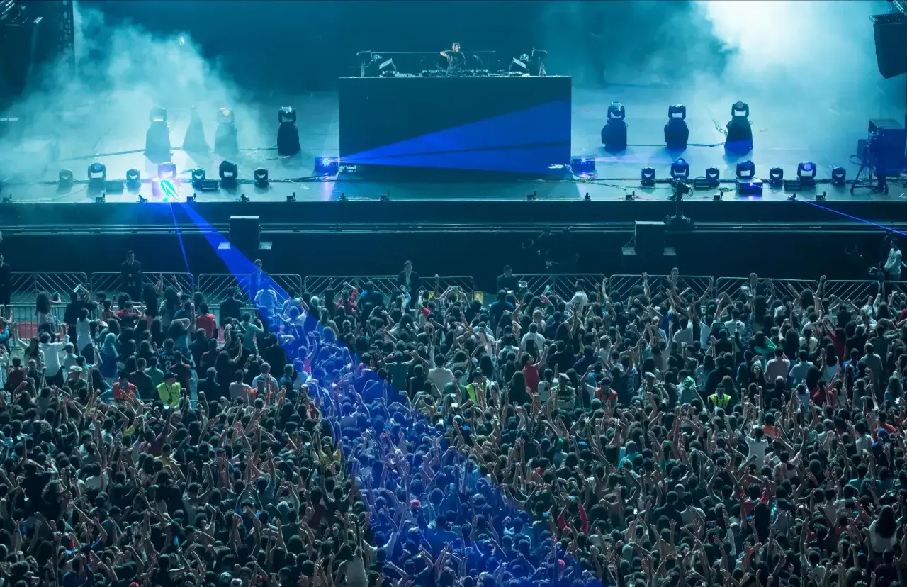 Tłum szaleje na koncercie, gdy DJ Avicii puszcza swoje hity. Niebieskie światła i dym tworzą niesamowitą atmosferę.