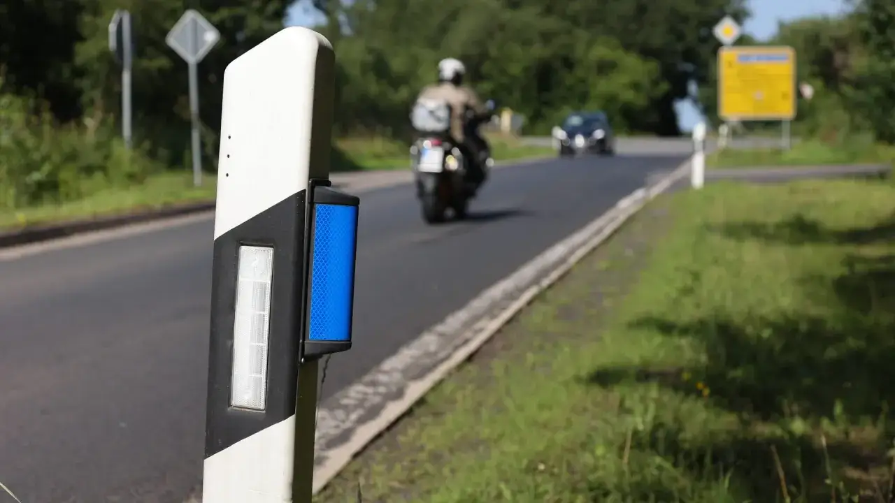 Pfosten mit wei&szlig;en und schwarzen Streifen, der blaue reflektoren f&uuml;r bessere Sichtbarkeit bei Nacht hat. Ein Motorradfahrer f&auml;hrt auf der Stra&szlig;e.