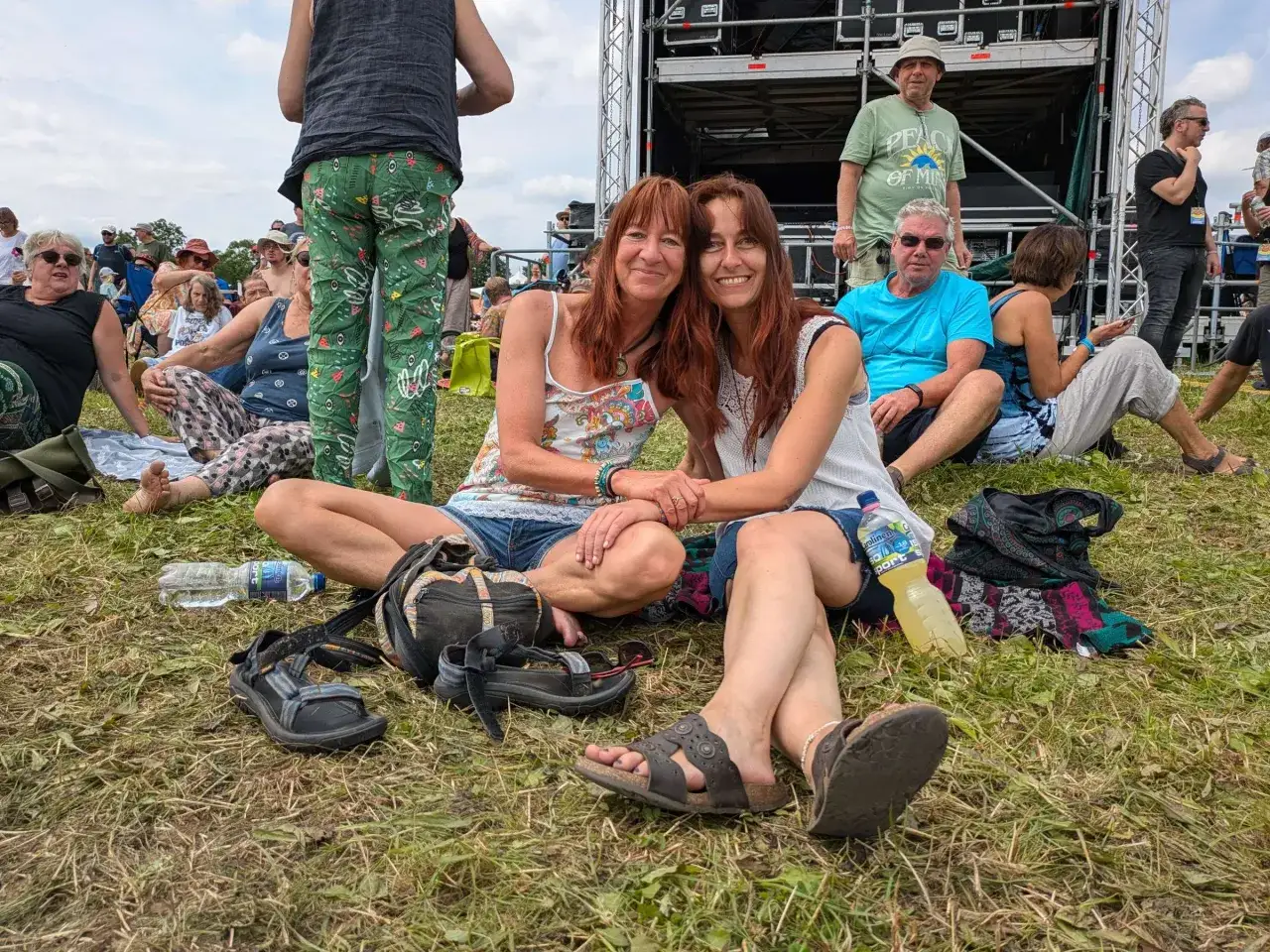 Zwei Frauen mit roten Haaren und lässiger Kleidung, die an einen Festival-Vibe erinnern, sitzen im Gras. Sie sind Teil einer Gruppe, die sich vor einer Bühne versammelt hat, und strahlen eine entspannte, neo-hippieske Atmosphäre aus.