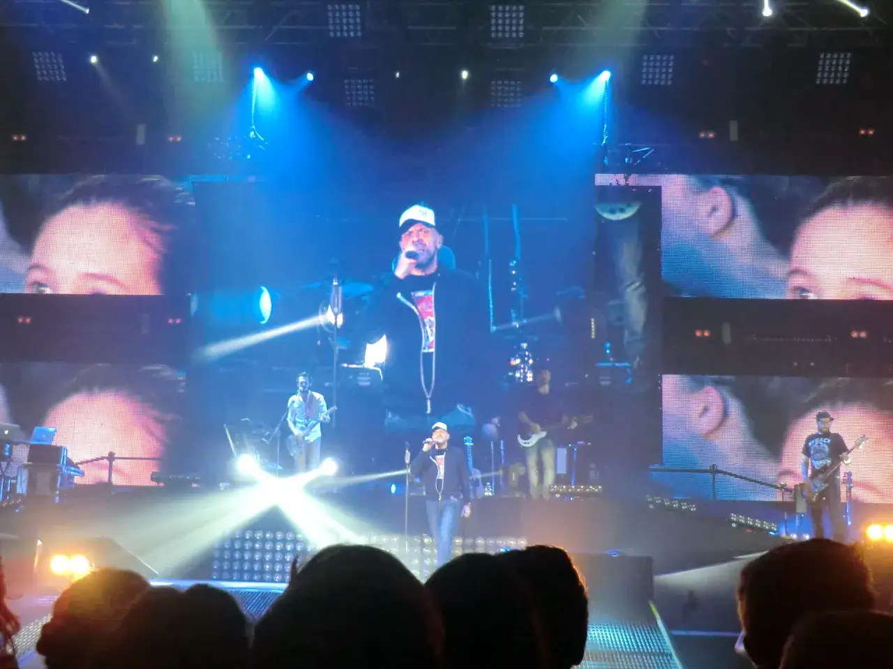 Sul palco, un cantante con cappellino bianco canta sotto i riflettori, mentre i musicisti suonano. Eccoti testo.