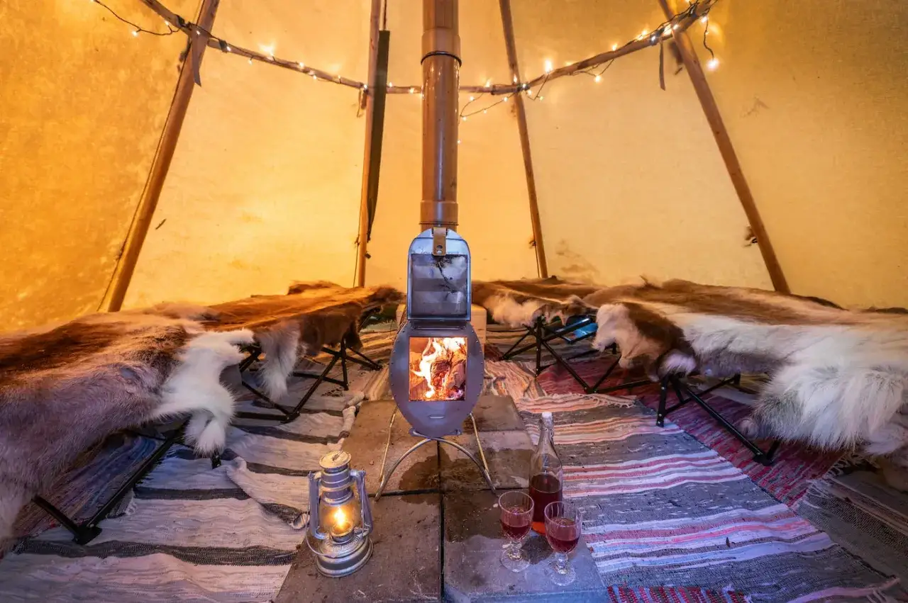 Mysig moskoselkåta med brasa, renskinn och tända ljus. En flaska och två glas står redo.