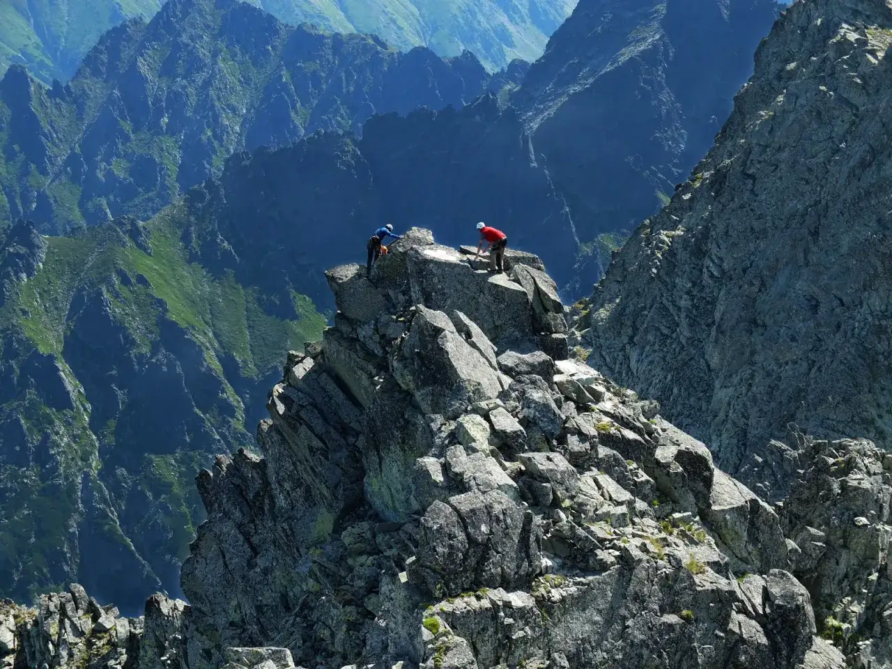 Dwóch alpinistów na skalistej grani podczas ekspozycji w górach. W tle piętrzą się kolejne, skaliste szczyty.