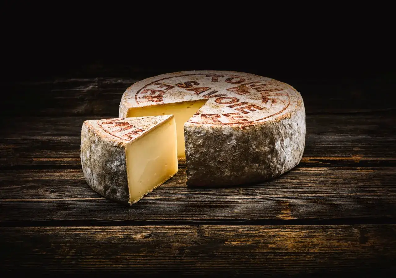 Une tome de Savoie, avec une part coupée, repose sur un fond de bois sombre.
