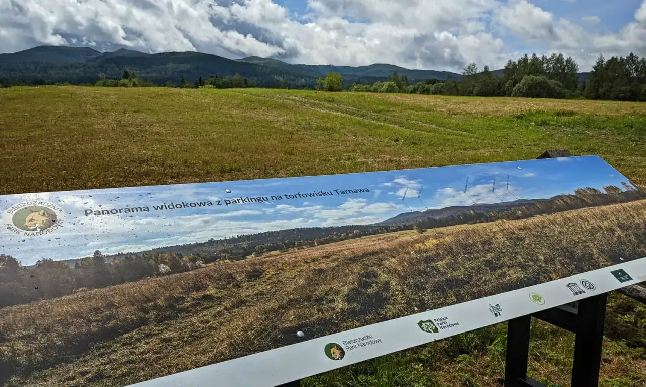 Tablica informacyjna z panoramą widokową na torfowisku Tarnawa. W tle góry i chmury.