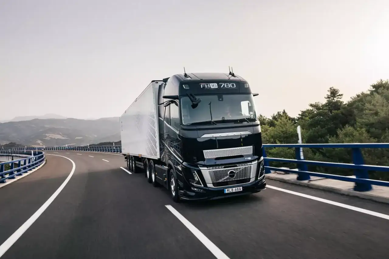 Czarny Volvo FH16 780 z białą naczepą jedzie po krętej drodze z widokiem na góry i lasy.