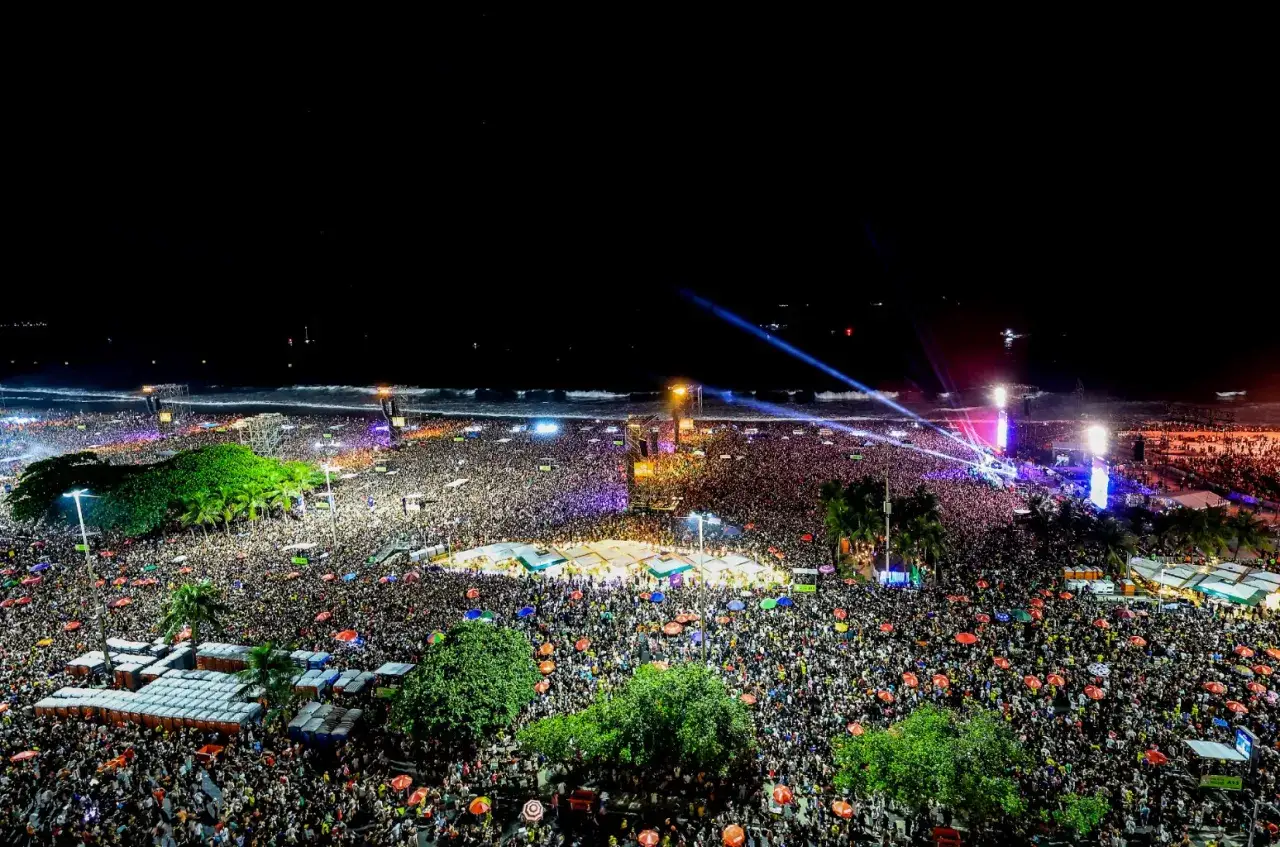 Nocny koncert Lady Gagi na plaży, tłumy ludzi pod sceną, fale oceanu i światła reflektorów tworzą niezapomniany widok.