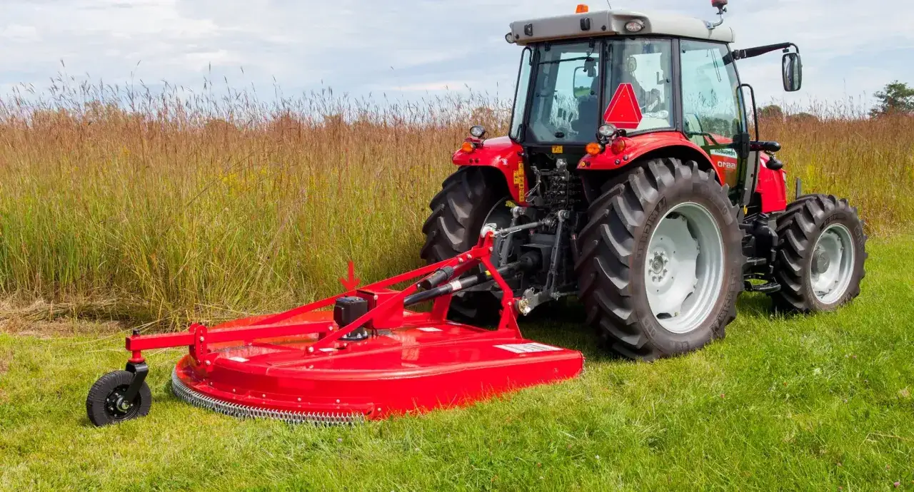 Czerwony traktor z zamontowaną kosiarką rotacyjną. Przekładnia kątowa WOM jest gotowa do pracy na polu.