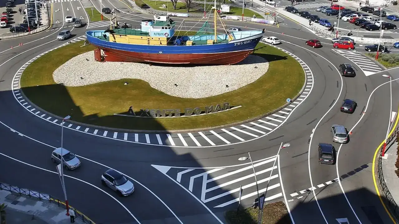 Un barco pesquero decorativo en el centro de una turbo rotonda. Coches circulan por la carretera.