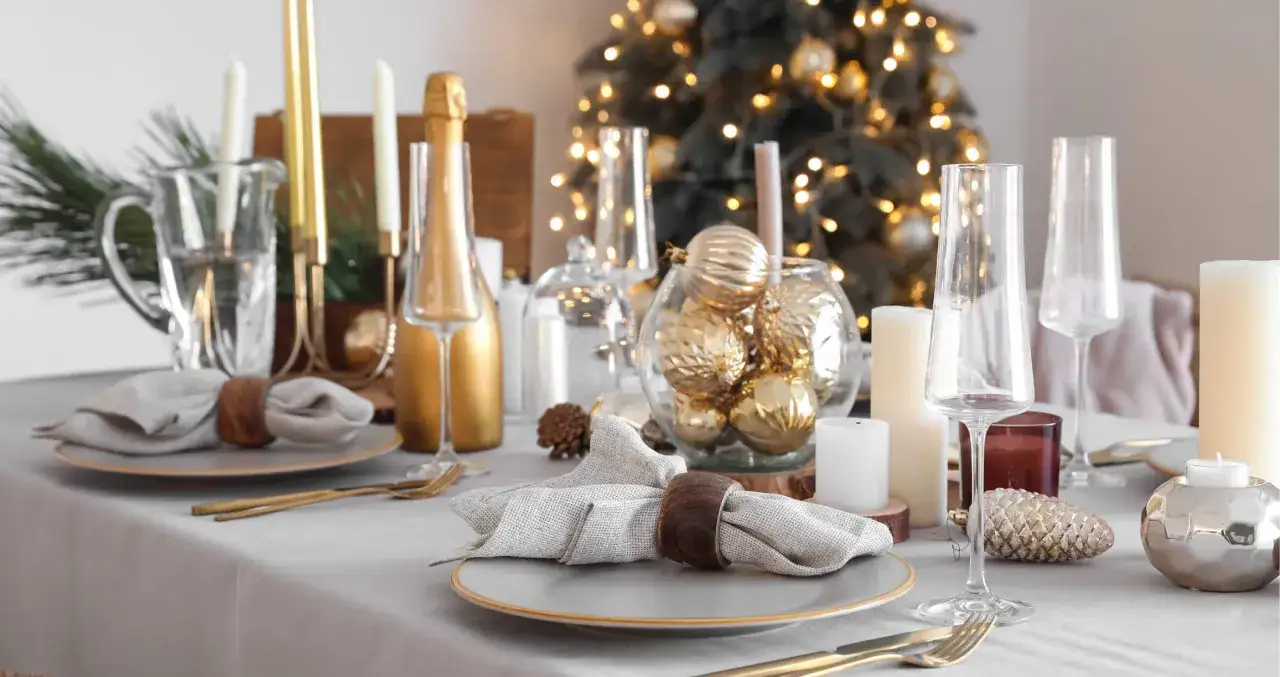 Moderne tischdeko weihnachten: goldene Kugeln, Kerzen und Champagnerflasche auf festlich gedecktem Tisch vor beleuchtetem Weihnachtsbaum.