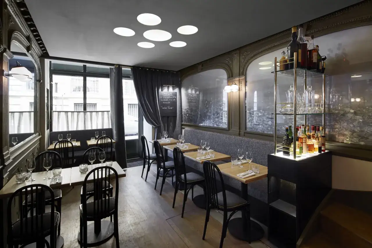 Przytulne wnętrze, co to jest bistro, z drewnianymi stołami, czarnymi krzesłami i półkami z butelkami alkoholu.