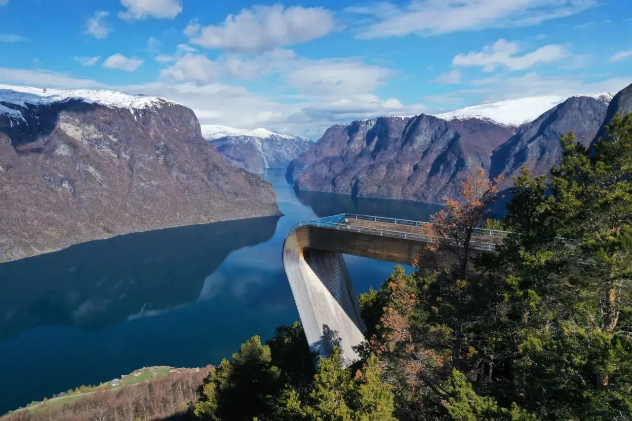 Spektakularne norwegia widoki: drewniana platforma widokowa nad fiordem, ośnieżone g&oacute;ry w tle, błękitne niebo z chmurami.