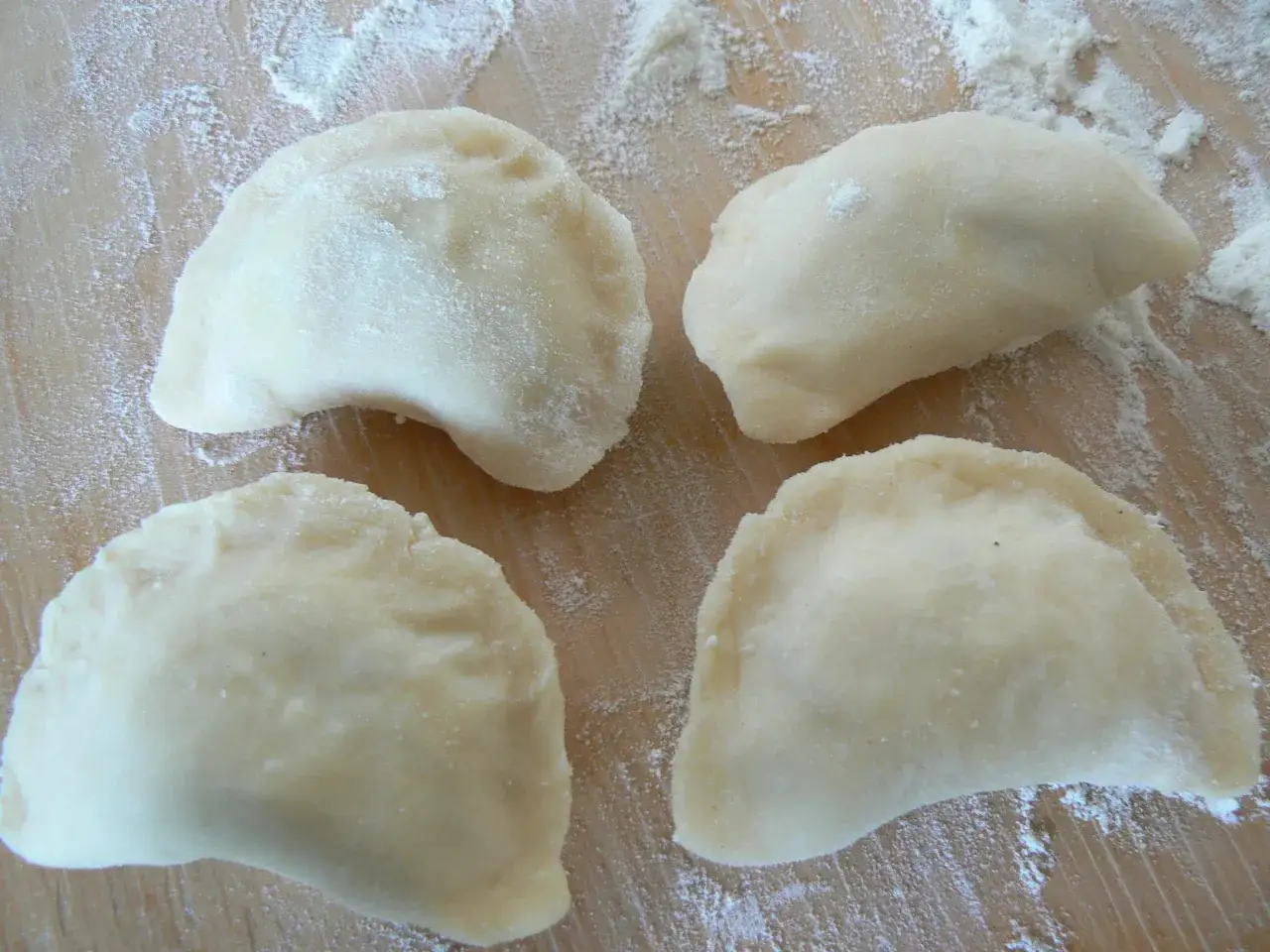 Cztery surowe pierogi leżą na drewnianej desce, posypanej mąką. To efekt udanego przepisu na miękkie ciasto na pierogi.