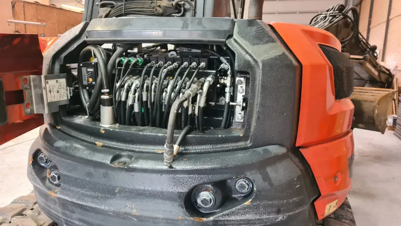 Widok na otwartą komorę hydrauliczną koparki. Wiele węży i zaworów. Zagęszczarka nie jedzie do przodu, prawdopodobnie problem z układem hydraulicznym.