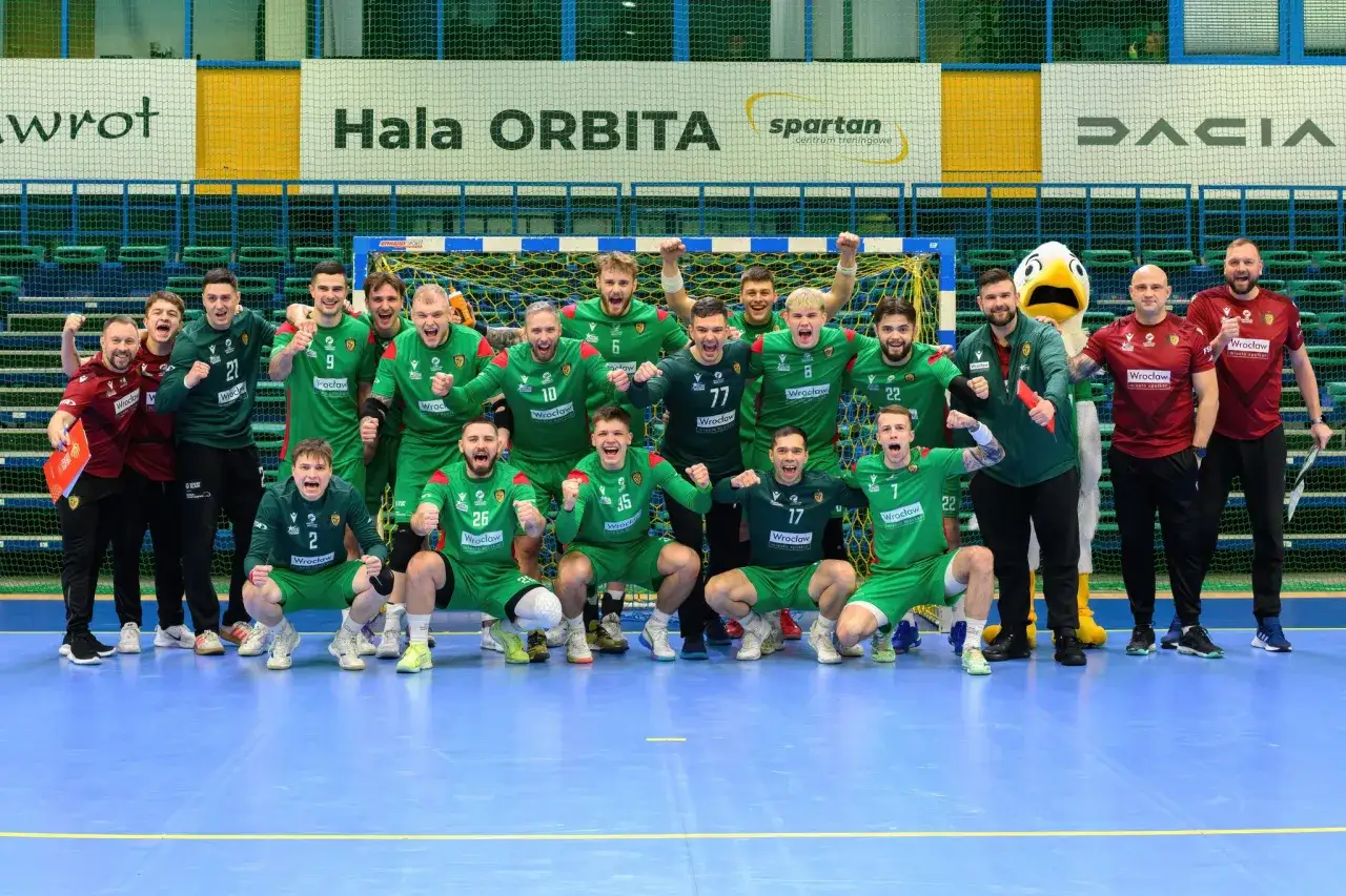 Drużyna piłki ręcznej w zielonych strojach świętuje zwycięstwo. Na tablicy wyników widnieje nazwa hali i sponsorzy.