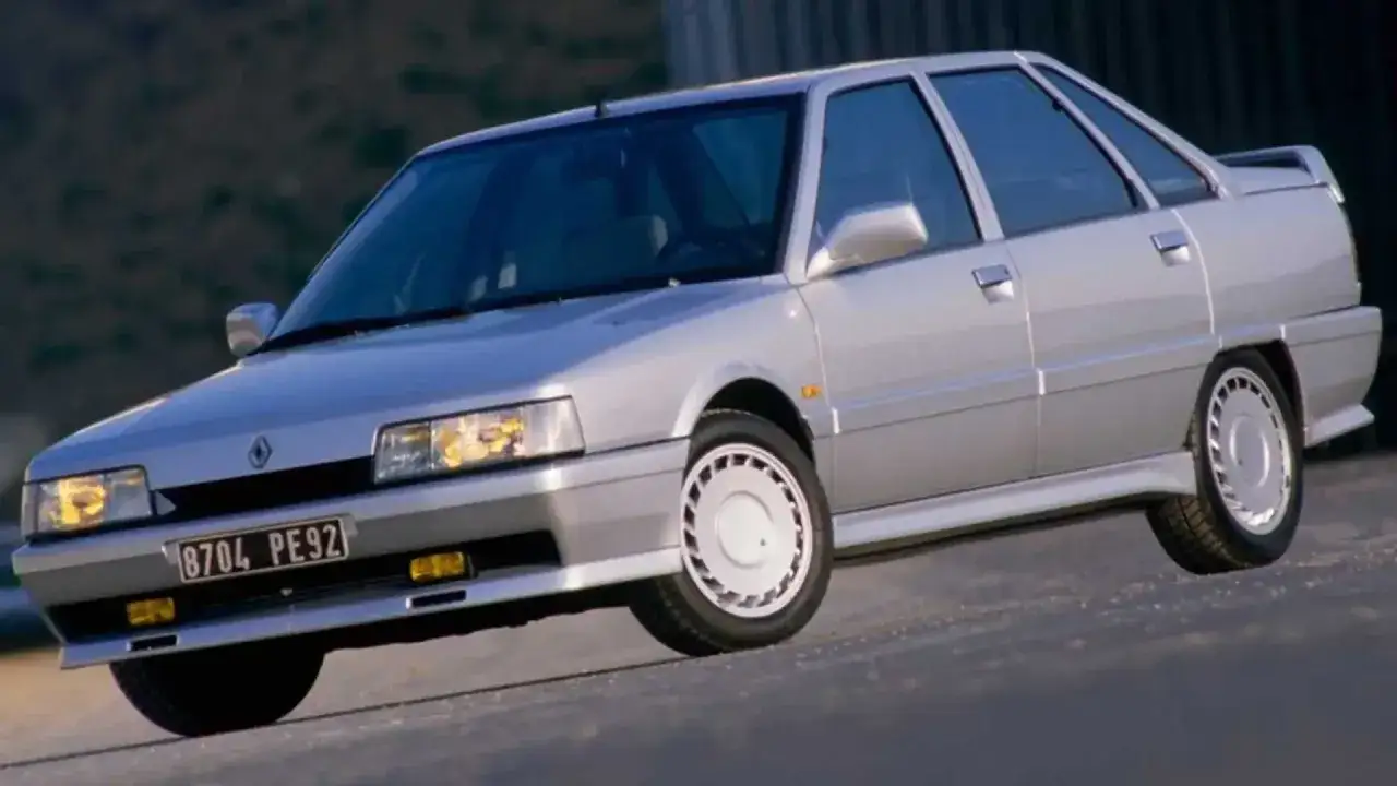 Un elegante Renault 21 Turbo plateado, un cl&aacute;sico de los 80, avanza por una carretera.