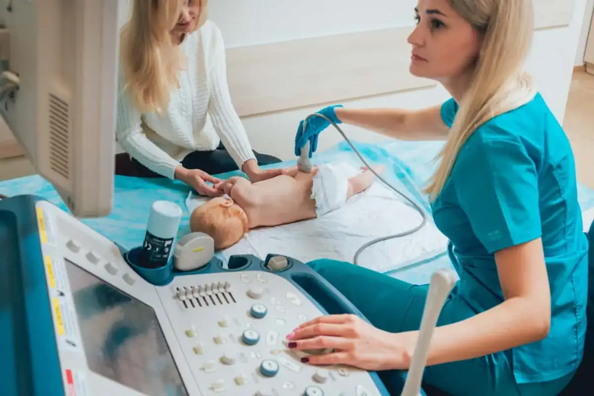Lekarka bada bioderka niemowlaka aparatem USG, aby wykluczyć dysplazję stawu biodrowego. Obok czuwa mama.