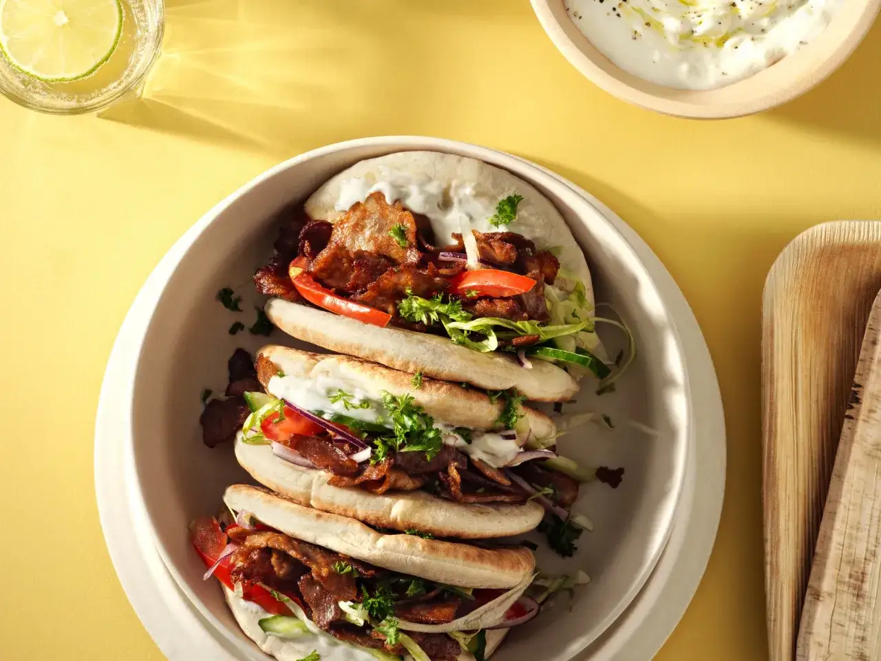 Tre gyros kebab-rullar fyllda med k&ouml;tt, gr&ouml;nsaker och s&aring;s. En glas med limevatten och en sk&aring;l med dipps&aring;s syns ocks&aring;.