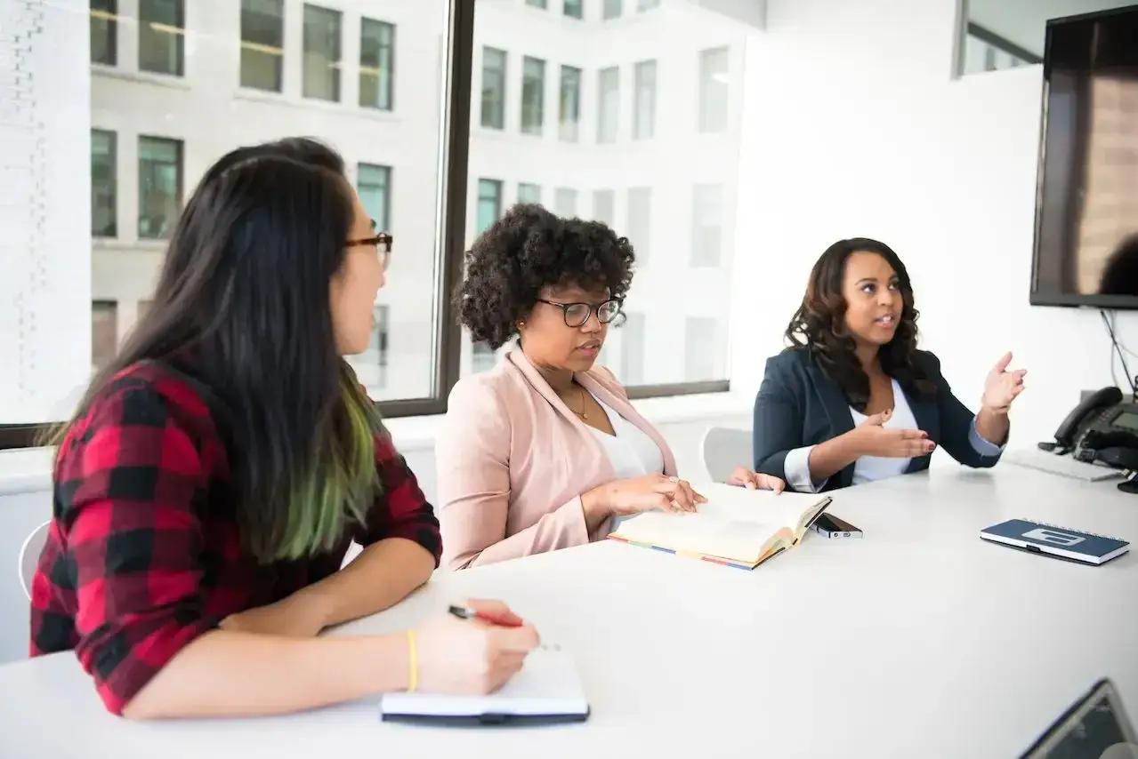 Tres mujeres en una reuni&oacute;n discuten ideas, cada una lista para aportar su perspectiva &uacute;nica.