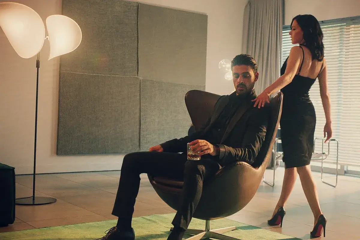 Elegancki mężczyzna w garniturze siedzi w fotelu z drinkiem, a kobieta w czarnej sukience stoi obok. Scena jak z filmu 