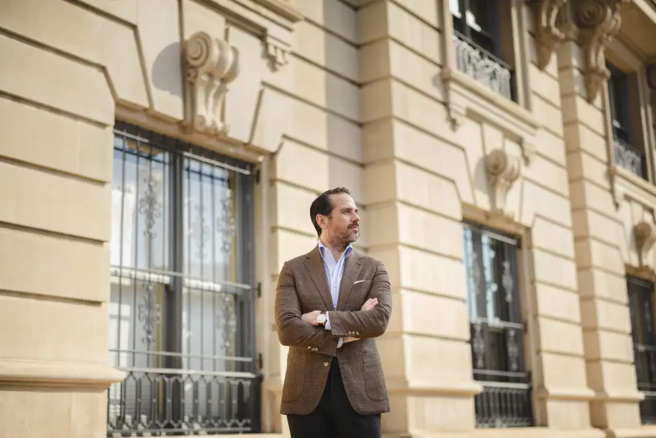 A man in a stylish brown sport coat outfit stands with arms crossed in front of a classic building.