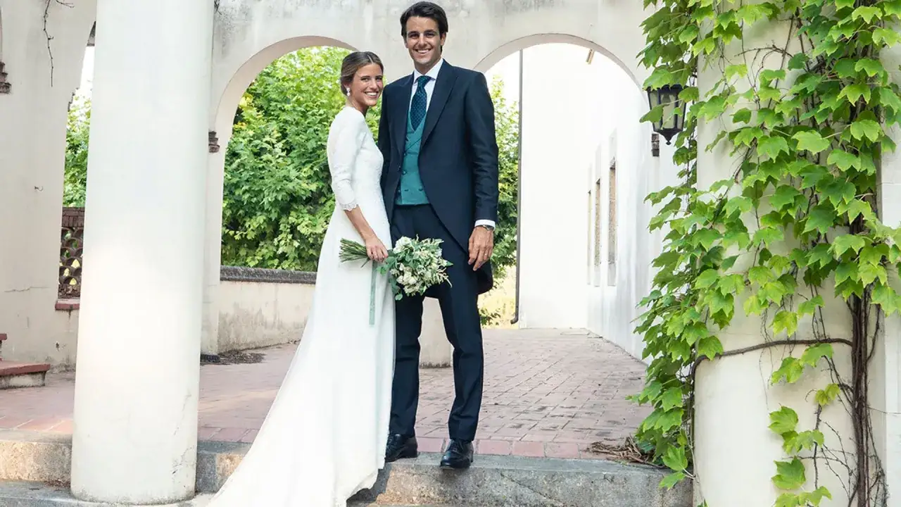 Novios sonriendo, ella con vestido blanco y ramo, &eacute;l con traje oscuro. Un escenario perfecto para bodas vintage.