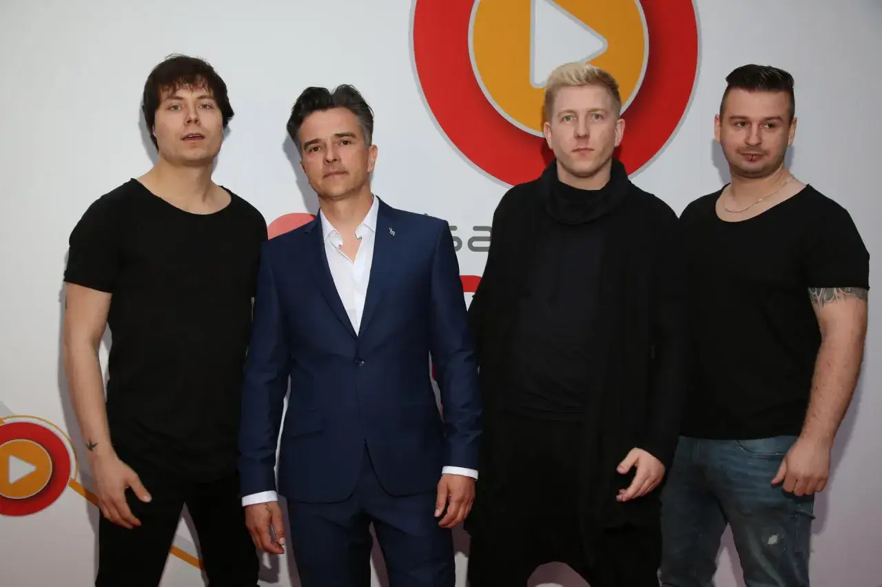 Czterech mężczyzn pozuje do zdjęcia. Dwóch w czarnych t-shirtach, jeden w granatowym garniturze, a czwarty w czarnej bluzie z kapturem. Wszyscy prezentują swoje **loka utwory**.